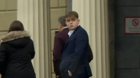 RTÉ A man is wearing a suit and standing outside a building with large white pillars. A do not smoke sign is on the wall. A woman and a man are walking in the background.