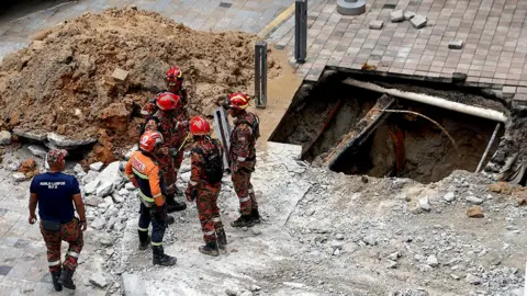 Rescue workers chat as they stand next to the sinkhole with a pile of rubble next to it, taken in Kuala Lumpur on 23 August