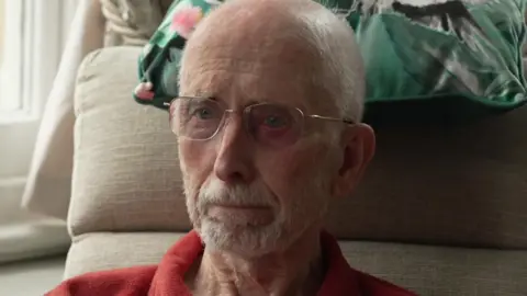 Brian Gundry, is a resident of a residential home in Dorchester. He is sittings in a chair in his room. There is a cushion behind his head. He's wearing a red shirt and has glasses and a beard.