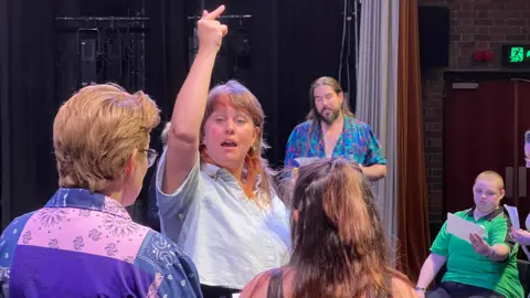 A woman is showing others what notes to hit by raising her hand as part of the rehearsals
