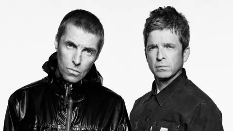 Liam and Noel Gallagher stand side by side in a black and white photo. Liam is wearing a dark zipped jacket and Noel is wearing a dark coloured denim jacket.