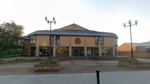 A modern, multi-story brick building with large windows and a central entrance. Two lampposts with multiple lights stand on either side of a paved area leading to the building.