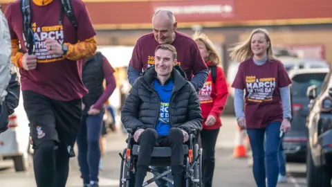 PA Media Stephen Darby is dressed in a blue "March Of The Day" t-shirt and an unzipped black coat. He smiles whilst being pushed in a wheelchair by another man dressed in a burgundy "March Of The Day" t-shirt.