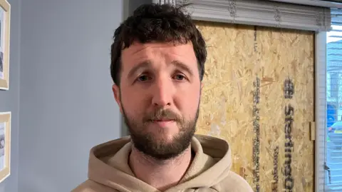Darian Moore is wearing a beige hoodie. He has short brown hair and and beard. He is standing near the boarded up window in his living room. 
