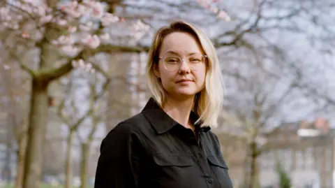 Mission 44 Cara Cinnamon is wearing glasses and a black button-up shirt, standing outdoors beneath blooming tree branches. The portrait is framed from the waist up, with a softly blurred background of trees and buildings in a park-like setting under natural daylight.