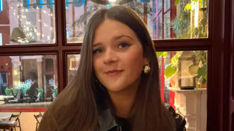 Stella Trimble looks into camera. She has a gold ear-ring and dark brown long hair. She is sitting in front of wooden framed window. Plants and candles can be seen behind with some reflective light shining back from one window pane.