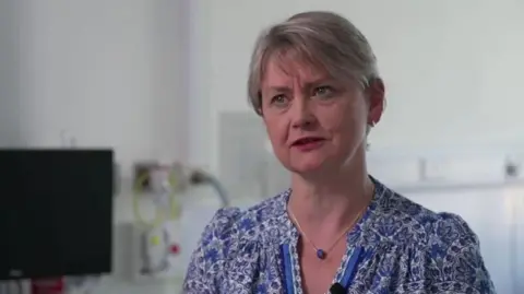 Yvette Cooper seated in a hospital clinical room with short hair, a necklace and patterned blue blouse. Medical equipment and a computer monitor are visible behind her against a white wall.