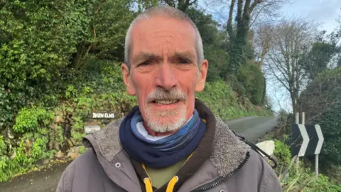Man with short grey hair and a white beard and moustache is looking at the camera. He is wearing a coat, with a brown fleece and yellow cord and has several neck scarves.