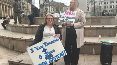 Two midwifery students holding signs and standing on steps in a square. One is looking at the camera and smiling and holding a placard which reads "3 years + 0 jobs = broken system". She is wearing a white top, has glasses and shoulder length blonde hair. The second woman is standing slightly behind her and looking to the left. She had a long brown cardigan on, black trousers and has blonde shoulder length hair. Her placard reads "NHS jobs, fairer bursaries, funded tuition".