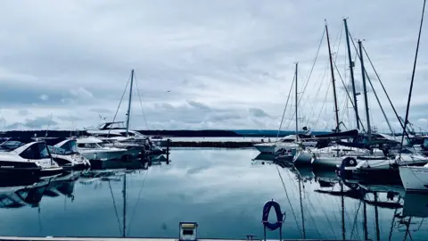 Dave boy Williams Two lines of moored yachts either side of the image sit on perfectly flat harbour waters which reflect the cloudy skies above. In the background breakwaters and the Dorset coastline 