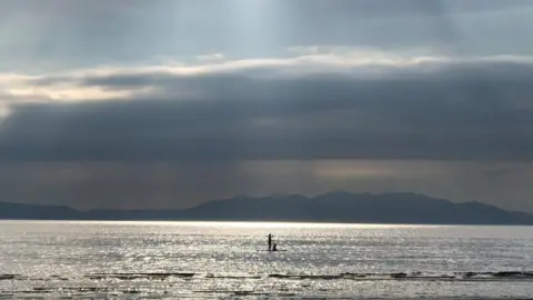 Carolyn Guild Lone paddleboarder crossing a sunlit sea beneath dramatic clouds.