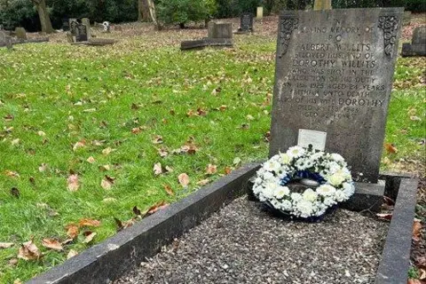 West Midlands Police A grave that is inscribed 'in loving memory of Albert Willits, beloved husband of Dorothy Willits, who was shot in the execution of his duty', with further words below. A wreath of white flowers is based at the foot of the grave, which is surrounded by grass.