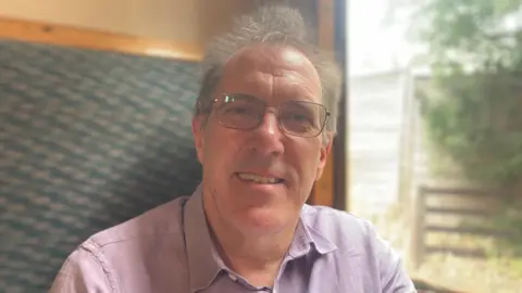Jenny Kirk/BBC Graham Hukins is in the middle of the image looking towards the camera with a light smile. He has short grey hair and is wearing black glasses and a purple shirt. He is sat on a seat in the train which is going past a wooden fence.