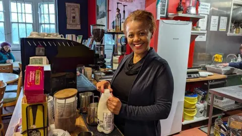 A woman wearing a black top smiling from behind a cafe counter. She's holding a bottle of milk and there is a coffee machine and a fridge behind her. A customer is sitting an one of the tables by a large window and the walls are painted red.