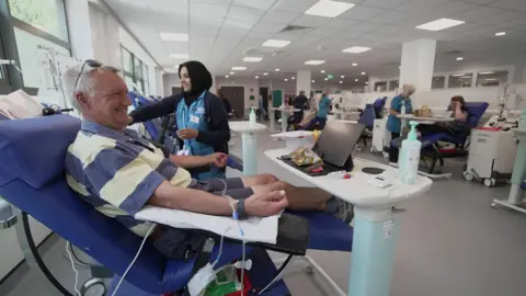 BBC A man smiles as he sits on a purple chair with a tube coming from his wrist as he donates blood.
