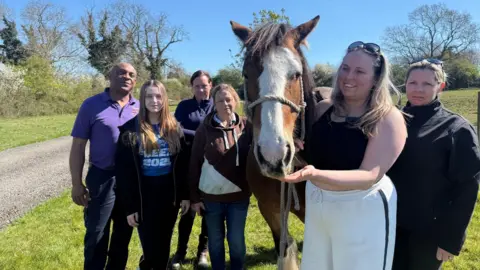 A man in a purple shirt stands alongside a five women and a horse with a white nose and brown fur in a green field. The horse is being held by a woman with blonde hair who is wearing sunglasses on the top of her head, a black top and white trousers.