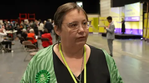 Esther Reeves wears a green stripy top and a black dress. A Green Party Rosette is pinned to her chest.