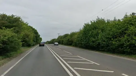 A general view of the A143 at Ixworth. Vehicles travel either side of the road. The road is lined with trees.