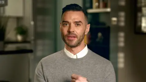 Matt Willis sits facing the camera - he has short black hair and wears a grey jumper