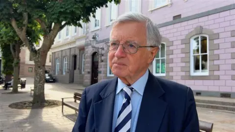 Deputy Sir Philip Bailhache wearing a blue suit and striped tie looks off to the left of the camera.