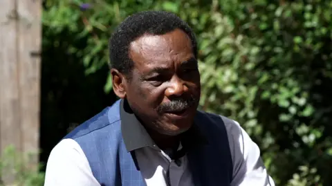 BBC/Steve Humphrey Close-up outdoor portrait of Sir Gordon Greenidge seated in a garden. He is wearing a light-colored shirt with a dark collar and a blue vest. Sunlight illuminates his face and clothing, while the background is softly blurred with green foliage and a wooden post visible on the left side.