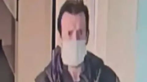 A CCTV image of Dean Hesketh, wearing a facemask and brown coat. It is of poor quality.
