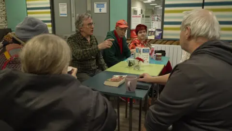 Steve Coogan is sat at a table talking with other people in the community centre