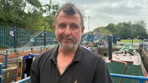 Dan Cauldwell, a man with stubble and wearing a black top, stands in front of some canal boats.