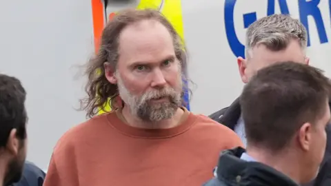 A man with long greying hair and a grey beard stands outside a garda van - he wears a red top