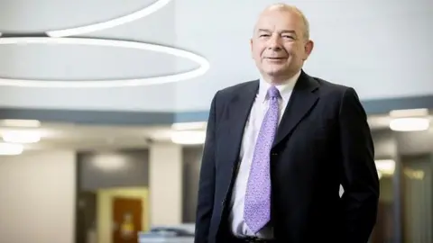 Coventry University A mostly-bald man wearing a suit jacket, white shirt and purple tie, stands in an office.