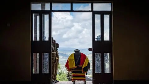 Badru Katumba/AFP Foto van de kist van de Oegandese atleet Rebecca Cheptegei in een hal met aan weerszijden soldaten, zodat mensen hun respect konden betuigen vóór haar begrafenis in het Bukwo-district, Oeganda - zaterdag 14 september 2024.