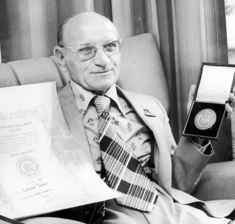 Getty Images A smiling Len Tasker wears a suit, patterned shirt and kipper tie. He is holding a certificate in one hand and a medal in the other. 