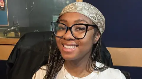 Handout Shaniyaa wears a white t-shirt and has large black framed glasses and has long brown braided hair. She wears a paisley patterened bandana on her head 