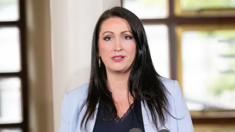 PA Emma is wearing a light blue blazer and navy vest top underneath.  She has long black hair and is wearing dangly gold earrings. 