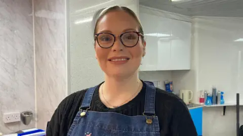 BBC Victoria Hoskins is wearing a blue apron, a black shirt and glasses. She is standing in her hair salon with white cupboards behind her and equipment on worktops.