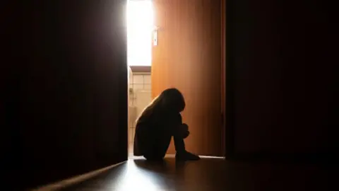 Despairing child in silhouette sitting with head on knees in the dark frame of a doorway, backlit by a room behind flooded with daylight - stock photo.