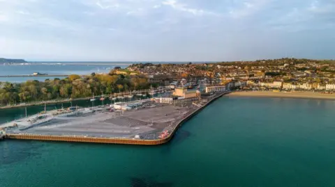 Dorset Council Aerial view, looking from the sea towards Weymouth Peninsula and Weymouth Beach. The peninsula is a flat triangle of land, mostly covered by a large car park. To the right of the peninsula is the sandy beach. To the left is the entrance to the harbour which is full of small boats on their moorings. On the far side of the harbour entrance is the tree-filled peninsula of Nothe Gardens.