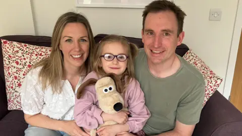 BBC Kate and Pete Hill sat on a sofa either side of their daughter Sofia who is holding a cuddly toy Gromit.