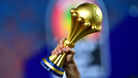 A hand holds up the gold Africa Cup of Nations trophy against a blurred background