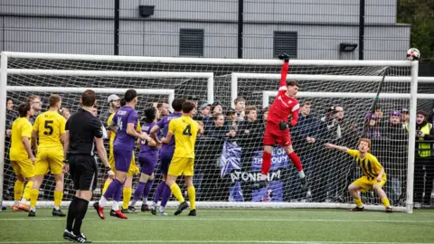 A group of footballers gathered around a goal as a ball comes flying in from the right. The goalkeeper, dressed in a red strip, is leaping into the air and holding the crossbar with one hand. 