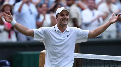 Arthur Fery of Britain celebrates winning his Men's Singles fourth round match with his arms stretched out in the air and smiling. He is dressed in a white polo shirt with a logo top right and a white hand and wrist sweatband. The crowd are cheering behind him. 