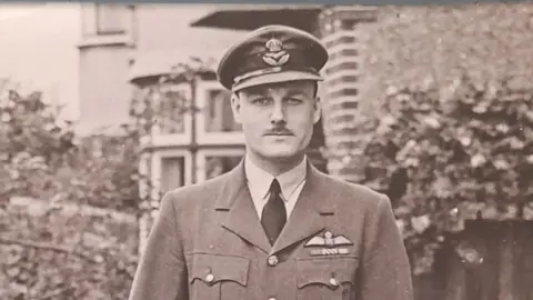 Norma Browne A black and white World War Two-era image of Group Captain Wilfred Surplice on the front cover of a self-published book by his son. He is wearing a Royal Air Force uniform, including a cap and jacket with RAF wings on them. He is in a garden with a wall, fence and house behind him.