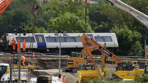 Eddie Mitchell Heavy machinery including cranes, excavators and construction vehicles is being used at the train derailment site near Lewes. A Southern train carriage is positioned beside the track while recovery crews work around the railway embankment and construction materials.