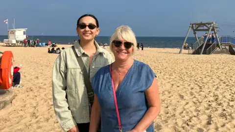 Lauren on the left and Caroline on the right standing on Bournemouth beach. Lauren has dark hair and is wearing a light coloured jacket and sunglasses. Caroline has blonde hair and is wearing a blue top and sunglasses.