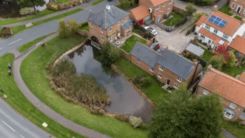 HSE An aerial shot of a pond surrounded by a green and a collection of houses