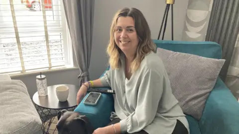 A woman is sat sideways in a deep blue chair, next to a small table, smiling at the camera. She is wearing a light grey-blue top and has a black dog sat at her feet. She has medium brown to fair shoulder length hair