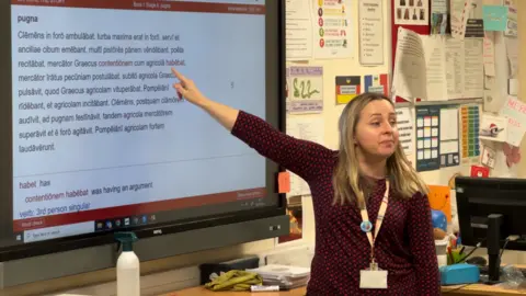 Teacher Marcella Jackson in a classroom. She is pointing behind her to a large TV screen containing Latin words. She has long blonde hair and wears a three-quarter length black top covered in a red spotted pattern and is looking out at the class. She wears a lanyard around her neck. 