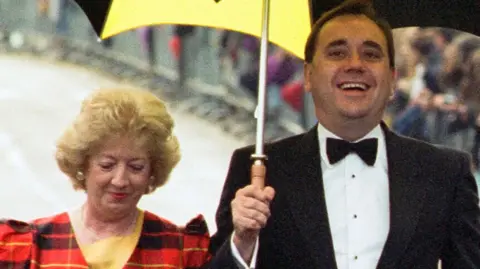 Alex Salmond with his wife Moira pictured at the Braveheart premiere in Stirling in 1995. Moira wears a tartan blazer while Alex wears a black suit jacket, white shirt and black bowtie. He is carrying a black and yellow umbrella.