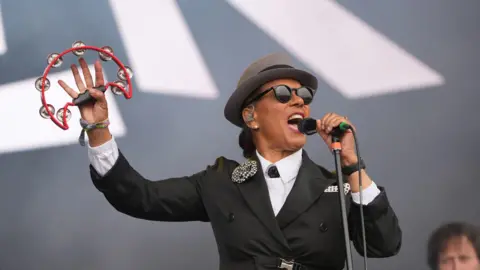 Pauline Black of The Selecter performing on stage while holding a tambourine. She wore all black, black sunglasses and a hat. 