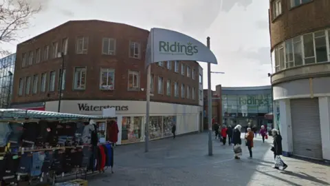 Buildings in an outdoor area with a white sign in the middle of the precinct with green writing on which says 'Ridings shopping centre'. There are a number of people pictured with shopping bags and a market stall in the foreground with clothes hanging on rails. 